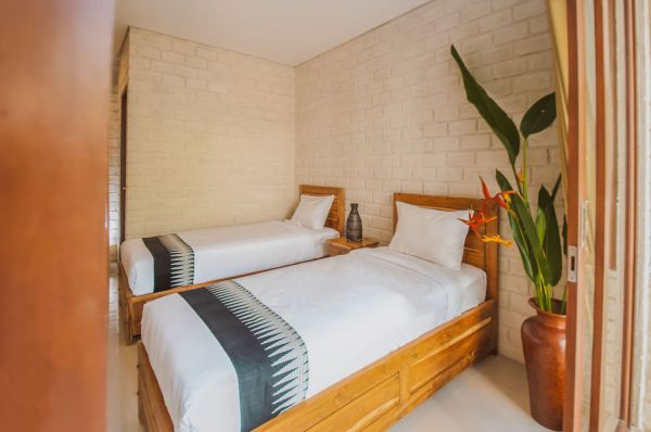 Cozy Balinese-style twin bedroom with wooden beds, white linens, tropical bird-of-paradise plant, and exposed brick walls.